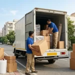 Professional packers and movers in Gurgaon safely loading cardboard boxes into a white moving truck in a residential neighborhood.